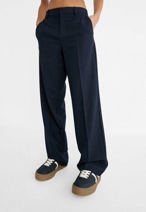Personne portant un pantalon large bleu marine foncé et des baskets noires avec des lacets blancs et des semelles marron, debout les mains dans les poches.