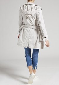 Persona caminando alejándose, vistiendo un abrigo trinchera con capucha en un beige claro, pantalones recortados de mezclilla azul rasgados y zapatillas blancas, sobre un fondo liso.