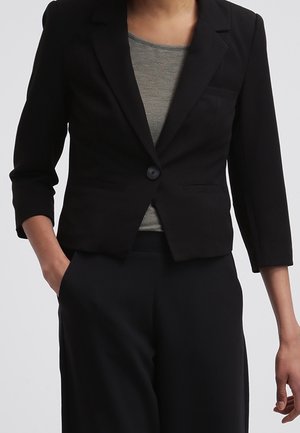 Femme portant un blazer noir court à manches trois-quarts sur un haut gris, associé à un pantalon noir taille haute.