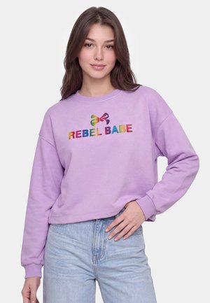 Ung kvinde med langt brunt hår iført en lilla sweatshirt med farverig "REBEL BABE" tekst og lyseblå jeans, poserer med den ene hånd i siden.