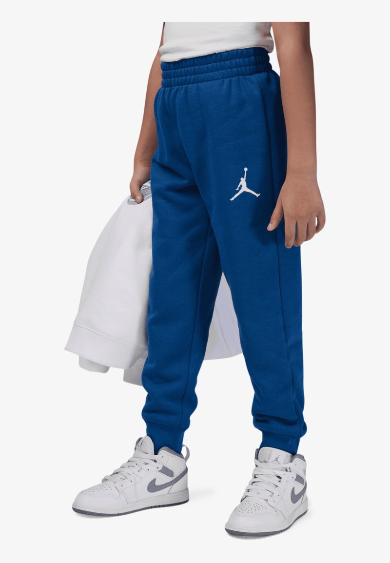 Pantalones de chándal azules de Jordan con cinturilla elástica, que presentan un logo Jumpman blanco. Llevados con un sudadera blanca y zapatillas Nike grises.