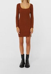 Robe en tricot côtelé marron à manches longues et décolleté carré, avec des fentes latérales. Assortie à des bottines noires.