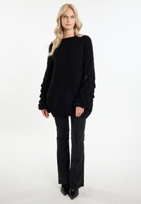 Maglione nero lavorato a maglia con maniche a sbuffo testurizzate. Abbinato a pantaloni neri a palazzo e tacchi a punta neri. Sfondo chiaro.
