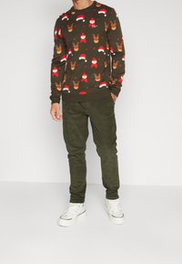 Mörkgrön tröja med röda mönster av jultomtar och renar. Matchad med olivgröna byxor och vita sneakers, vilket ger en avslappnad look.