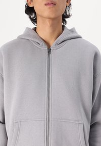 Γκρι ζακέτα με φερμουάρ κατασκευασμένη από μαλακό ύφασμα, που διαθέτει κουκούλα με κορδόνι, μπροστινές τσέπες και ομαλή κλεισίματος φερμουάρ. Ορατές λεπτομέρειες με υφή στην επιφάνεια.