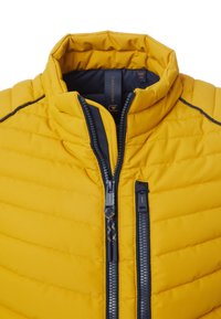 Gelbe Daunenjacke mit hohem Kragen, ausgestattet mit einem Reißverschluss vorne und einem kontrastierenden schwarzen Reißverschluss. Strukturierter, gesteppter Stoff mit horizontalen Nähten.