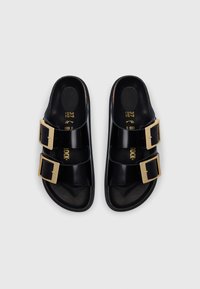 Μαύρα πλαστικά σανδάλια με δύο φαρδιές λουριά που διαθέτουν χρυσές τετράγωνες αγκράφες. Λείο υλικό, επίπεδες σόλες και σχεδιασμός σόλας με ανάγλυφη μορφή. Μέγεθος 37.