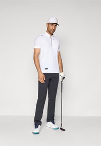 Biała koszulka polo z krótkim rękawem i zamkiem przy kołnierzyku, ciemnoszare spodnie, białe buty golfowe z niebieskimi akcentami oraz biała czapka, trzymająca kij golfowy.