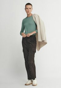 Hellgrüner langärmeliger Pullover kombiniert mit dunkelbraunen Cargohosen, die Taschen und eine lockere Passform bieten. Weiße Sneakers und ein beigefarbener Mantel, lässig über die Schulter geworfen.