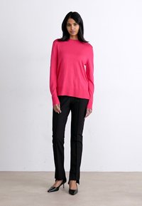 Roza svetlo roza pulover, rebrast izrez in manšete, oblečen z črnimi ozkimi hlačami in črnimi visokimi petami, stoječa na nevtralni ozadju.