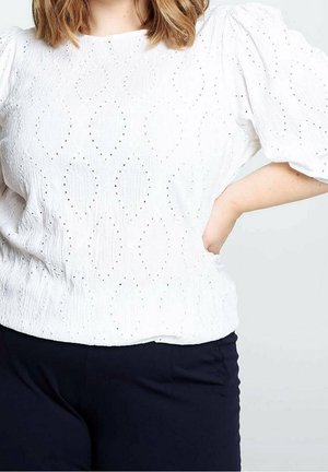 Femme aux cheveux mi-longs portant un chemisier blanc texturé à manches ballon et un pantalon noir, main posée sur la hanche.