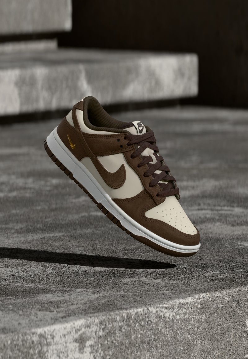 Καφέ και μπεζ αθλητικό παπούτσι Nike με κορδόνια, στον αέρα πάνω από σκάλες από σκυρόδεμα, ρίχνοντας σκιά από κάτω.