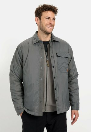 Gray, quilted jacket with a button closure, chest pocket, and a smooth, shiny texture. Worn over a gray knit sweater and black pants.