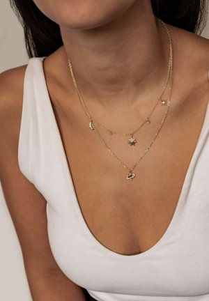 Zwei geschichtete Goldketten mit himmlischen Anhängern, darunter eine Sonne, ein Halbmond und kleine Sternanhänger, getragen mit einem weißen ärmellosen Oberteil.