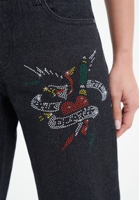 Black denim jeans featuring a rhinestone design with a heart, snakes, and decorative ribbons in red, white, green, and yellow accents.
