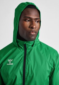 Mężczyzna ubrany w jaskrawozieloną kurtkę z kapturem, z czarnym zamkiem i białym logo hummel, patrzący na bok na jednolitym szarym tle.