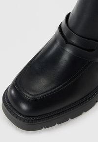 Stivale alla caviglia in pelle nera con finitura liscia, punta arrotondata e dettagli cuciti lungo la suola. Presenta una caratteristica fascia sulla parte superiore.