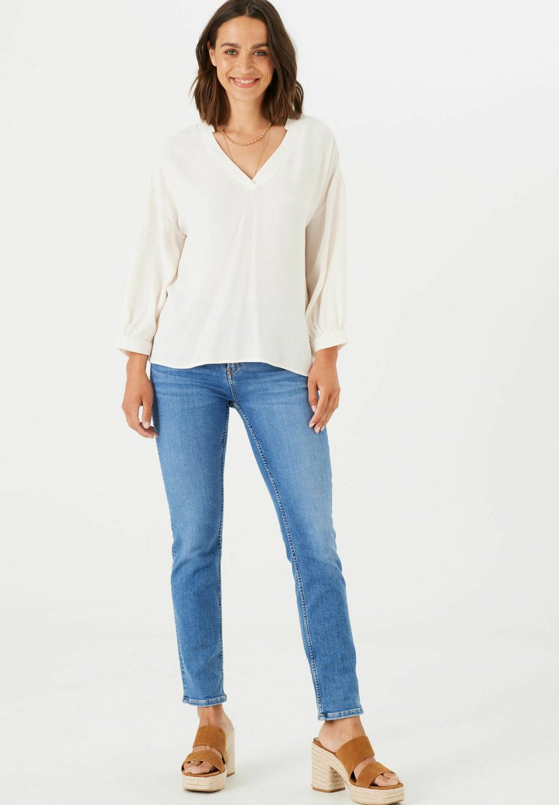 Blusa blanca de cuello en V con mangas abullonadas; combinada con jeans ajustados azules y sandalias de plataforma marrones. Tejido suave con un ajuste relajado.