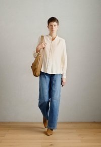 Camicia button-up beige, jeans baggy blu chiaro, mule con tacco color sabbia e borsa a tracolla in pelle marrone, su uno sfondo neutro.
