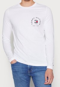 T-shirt bianca a maniche lunghe in cotone, con scollatura tonda e un logo circolare nero con la scritta “TOMMY HILFIGER EST. 1985” sul petto.