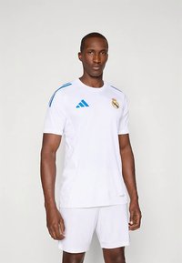Tricou Adidas alb cu mânecă scurtă, accente albastre și sigla Real Madrid, asortat cu pantaloni scurți albi. Fabricat din material ușor.