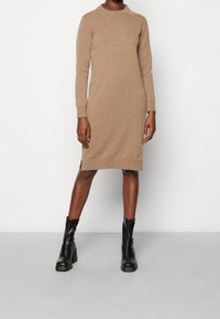 Robe en maille marron à manches longues et col rond, avec une fente latérale à l'ourlet. Associée à des bottines noires.