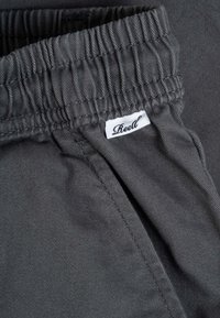 Pantalones con cintura elástica de color gris oscuro, costuras visibles y una pequeña etiqueta de tela blanca con la marca "Reell" cerca del bolsillo.