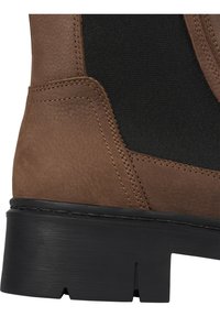Brown faux leather ankle boot with elastic side panels, textured surface, black rubber sole, and visible stitching details.