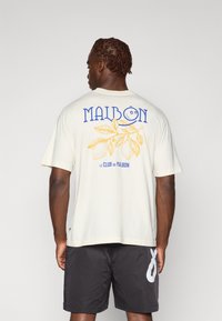 Lys kremfarget t-skjorte med blå og gul grafisk design på baksiden, inkludert tekst og et sitronmotiv. Kombinert med mørke shorts.