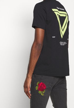 Osoba ubrana w czarny t-shirt z geometrycznym wzorem i napisem na plecach oraz czarne dżinsy z czerwoną haftowaną różą na uda.