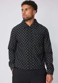 Homme avec des cheveux courts et bouclés portant une veste noire à boutons avec un motif en chaîne gris clair, se tenant avec les mains dans les poches.