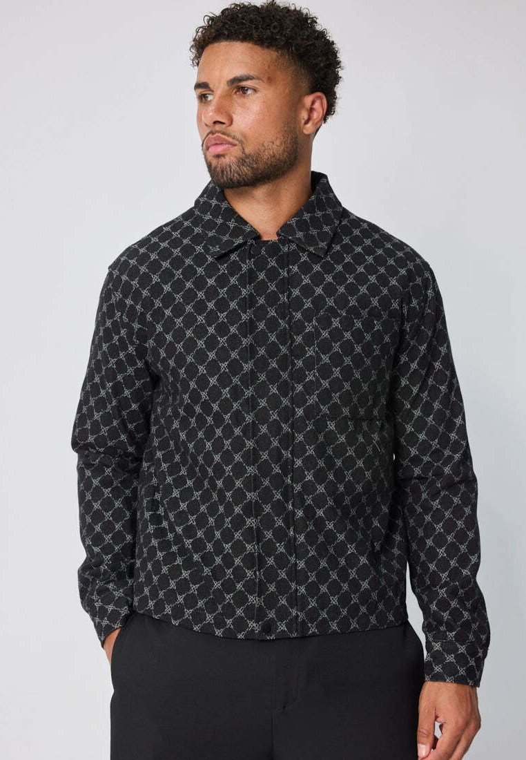 Homme avec des cheveux courts et bouclés portant une veste noire à boutons avec un motif en chaîne gris clair, se tenant avec les mains dans les poches.