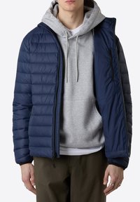 Giubbotto imbottito blu navy con design a trapunte, cappuccio e tasca sul petto con zip, indossato sopra un felpa grigia e pantaloni verde oliva.