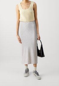 Cremfarbener, gerippter Tanktop mit gewellten Kanten, kombiniert mit einem hellgrauen, gerippten Midirock und grauen Sneakers, hält eine schwarze Handtasche.