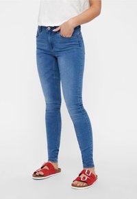 Blaue enge Jeans mit hoher Taille, ausgestattet mit zwei vorderen Taschen und einer glatten Textur. Getragen mit roten Rutsch-Sandalen.