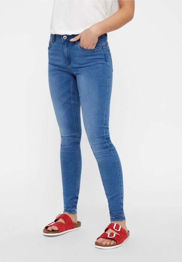 Blaue enge Jeans mit hoher Taille, ausgestattet mit zwei vorderen Taschen und einer glatten Textur. Getragen mit roten Rutsch-Sandalen.