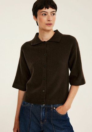 Vrouw met kort donker haar die een donkerbruine, geknoopte cardigan met ellebooglange mouwen en blauwe spijkerbroek draagt, staat met één hand in haar zak.