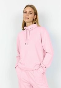 Žena nosící světle růžovou mikinu s kapucí na stažení a růžové kalhoty, stojící s rukama v kapse před jednobarevným pozadím.