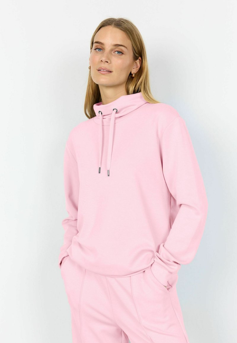 Žena nosící světle růžovou mikinu s kapucí na stažení a růžové kalhoty, stojící s rukama v kapse před jednobarevným pozadím.