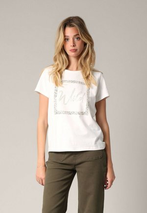 T-shirt en coton blanc avec un graphique argenté pailleté "Wild" dans un bord rectangulaire, associé à un pantalon vert olive.