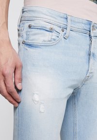 Jeans in denim azzurro chiaro con un design strappato, caratterizzati da due strappi visibili, cinque tasche e dettagli in metallo per chiusure e accenti.