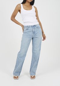 Kobieta w białym topie bez rękawów, jasnoniebieskich dżinsach o wysokim stanie, białych sandałach i srebrnej bransoletce, pozująca na tle jednolitego tła.