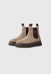 Beige ruskind Chelsea støvler med mørkebrune elastiske sider, trækkroge, tykke sorte såler og en subtil mærke tæ label på en hvid baggrund.