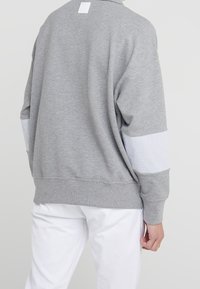 Persona vistiendo una sudadera gris con coderas blancas y pantalones blancos, vista desde atrás contra un fondo neutro.