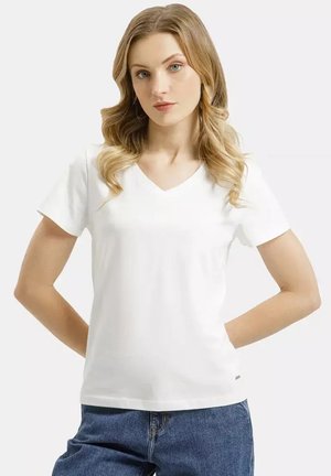 Młoda kobieta o blond włosach ubrana w prostą białą koszulkę z dekoltem w serek i niebieskie dżinsy, stojąca z rękami za plecami.