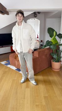 Veste en polaire blanche avec une poche frontale, jeans gris clair et baskets argentées. L'arrière-plan comprend une plante verte et un canapé à rayures brunes.