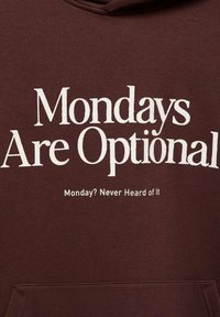 Hnědá mikina s kapucí a bílým textem "Pondělky jsou volitelné" a "Pondělí? Nikdy jsem o něm neslyšel." Tkanina má hladkou strukturu.