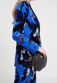 Černá kulatá kabelka se zlatým zipem, logem a řetízkovým popruhem, doplněná modrým sametovým sakem s květinovým potiskem.