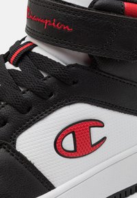 Sorte og hvite sneakers med teksturert læroverdel, røde detaljer, svarte snøring og gummisåle med en fremtredende logo.