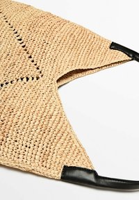 Torebka z plecionej słomy w naturalnym kolorze z czarnymi skórzanymi akcentami; ma trójkątny kształt i teksturowaną powierzchnię.